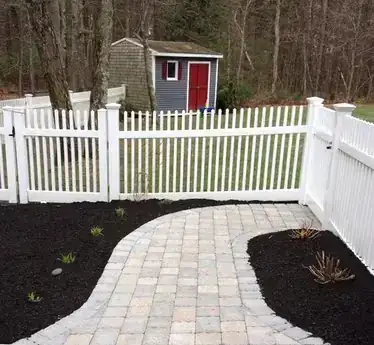 A white picket fence beside a paver walkway and garden beds